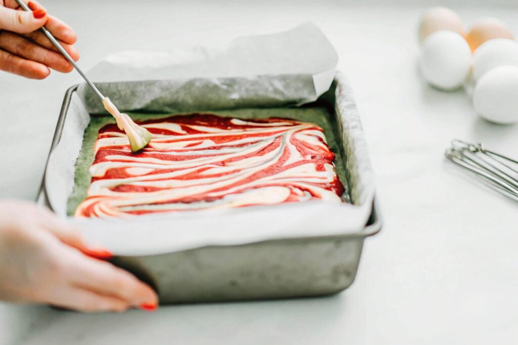 A baker using a skewer to swirl cream cheese into vibrant red batter in a parchment-lined pan.