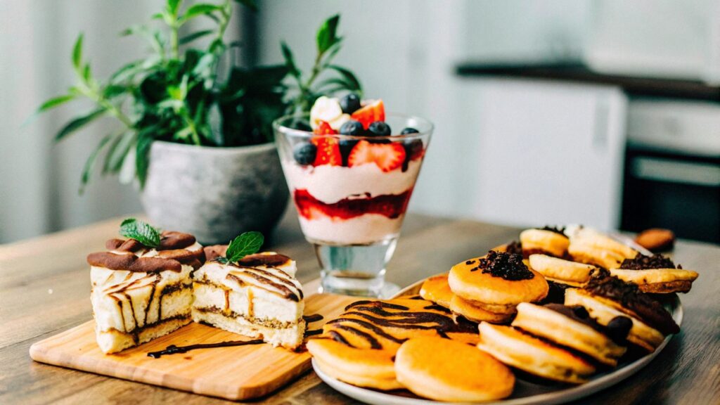 A dessert spread showcasing cottage cheese-based treats: a layered berry parfait, a cheesecake slice with fresh fruit, chocolate mousse with chocolate shavings, and golden pancakes drizzled with syrup, arranged on a rustic wooden table with a cozy kitchen backdrop