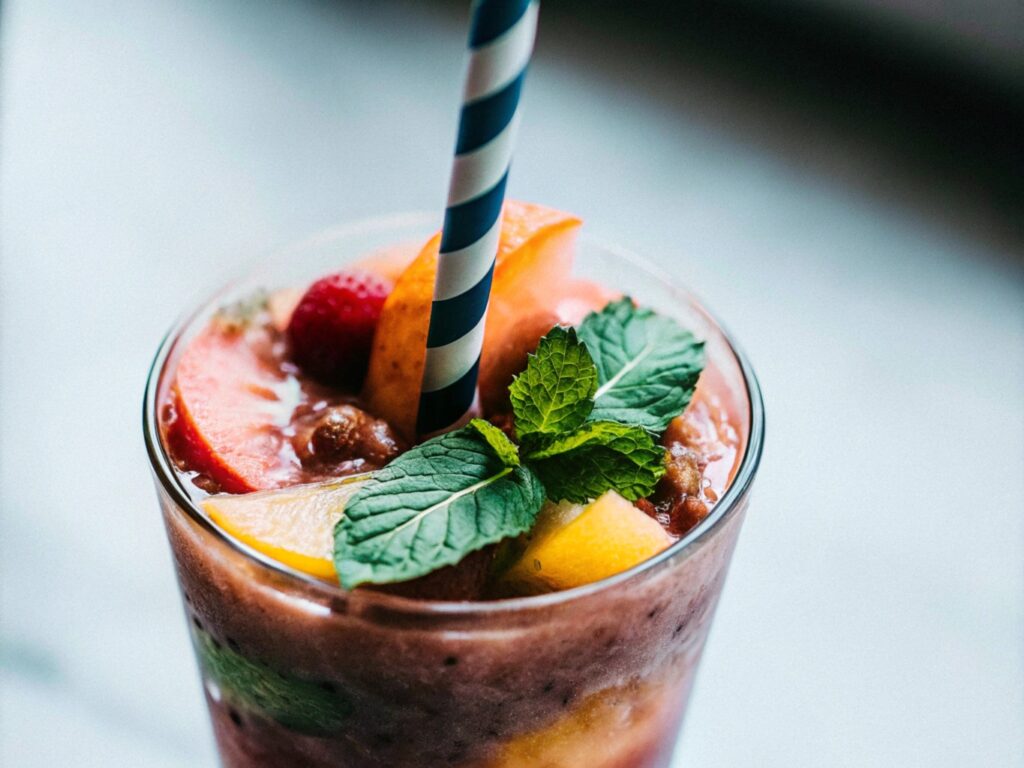 A colorful slushie in a clear glass, layered with bright red, blue, and green, topped with whipped cream and a striped straw on a sunny table