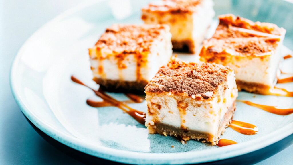 A close-up of churro cheesecake squares on a serving plate, dusted with cinnamon sugar and garnished with a drizzle of caramel sauce