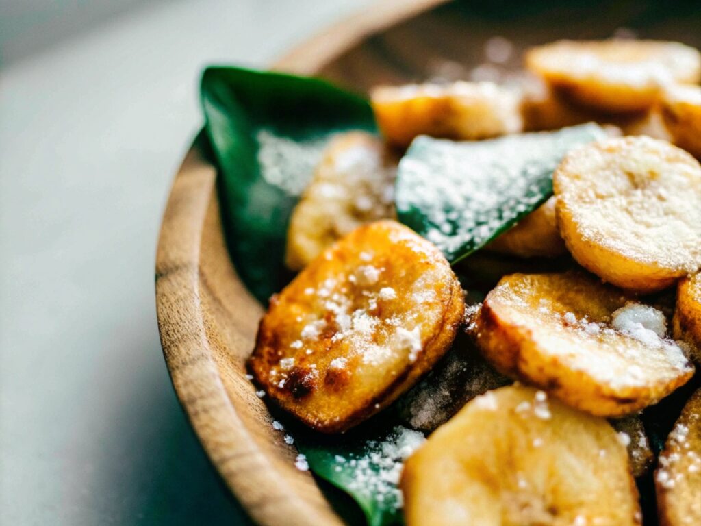 Close-up of golden fried banana slices garnished with powdered sugar, served on a rustic wooden plate.