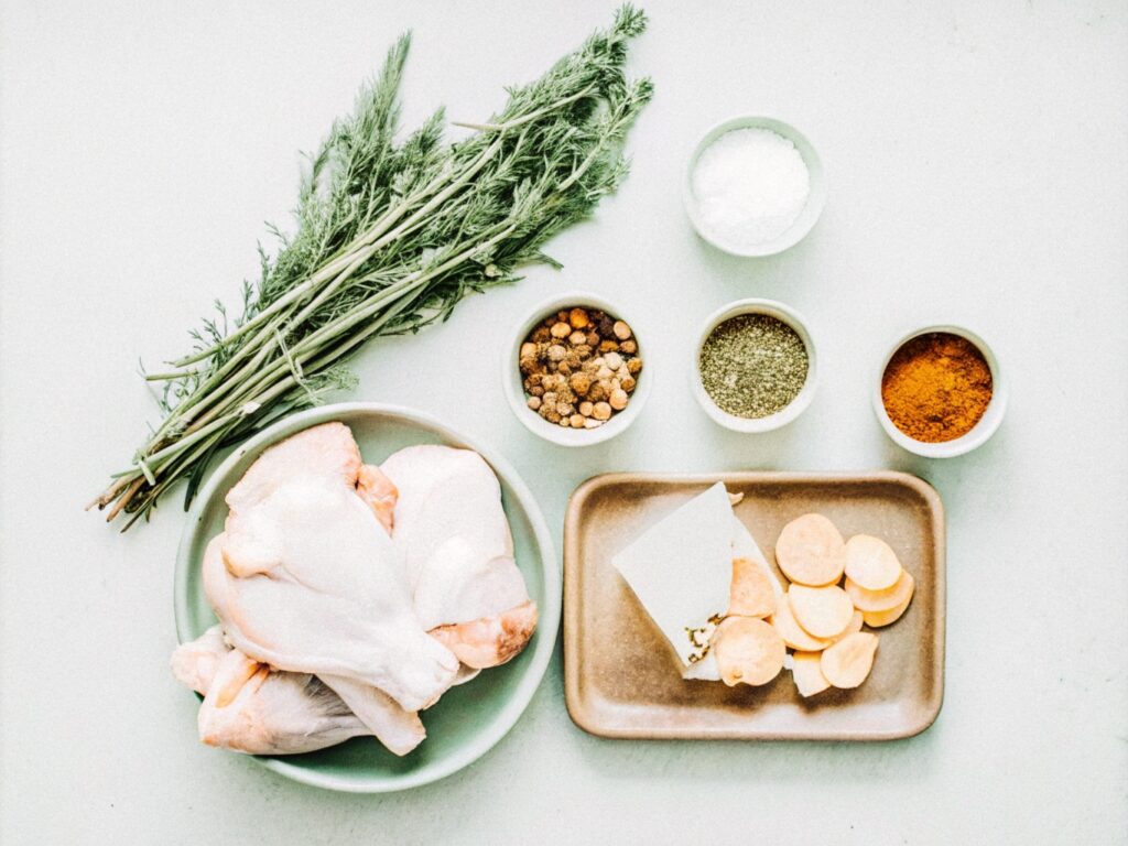 A flat lay of ingredients for smoked chicken thighs, including raw chicken, small bowls of spices, brown sugar, olive oil, and wood chips, arranged on a wooden surface.