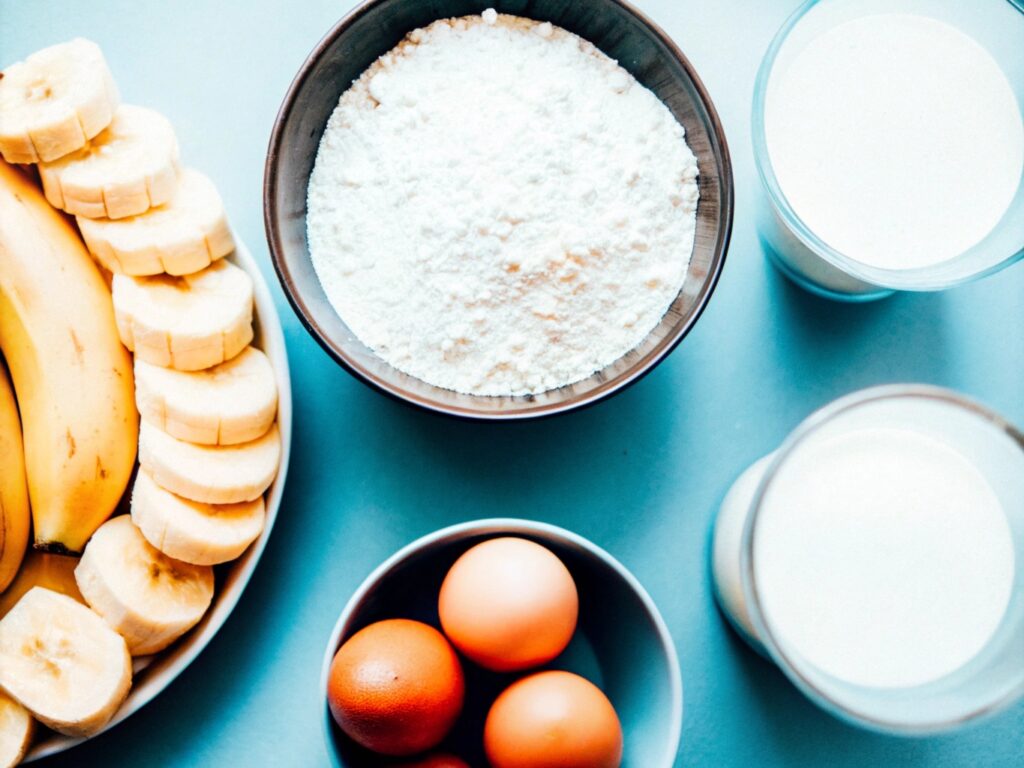 A flat lay of ingredients, including bananas, flour, milk, and eggs, arranged neatly on a kitchen counter with measuring cups and utensils