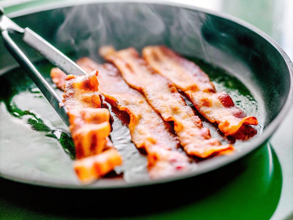 Strips of bacon sizzling in a skillet with steam rising, cooked to a crispy texture