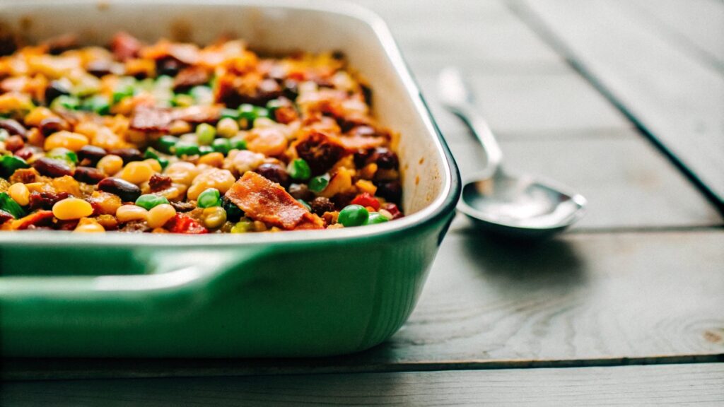 A vibrant casserole dish of Calico beans with layers of colorful beans, ground beef, and crispy bacon, served on a rustic wooden table with a serving spoon nearby.