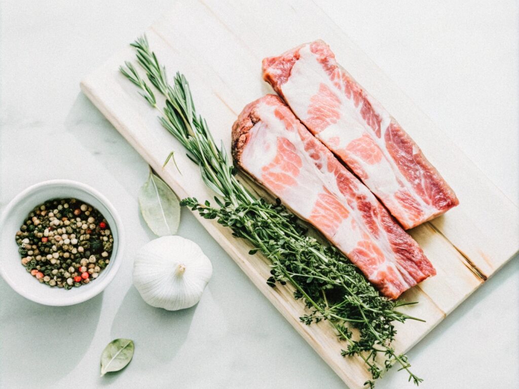 Raw boneless short ribs on a wooden cutting board with fresh herbs, garlic cloves, and a bowl of spices ready for preparation