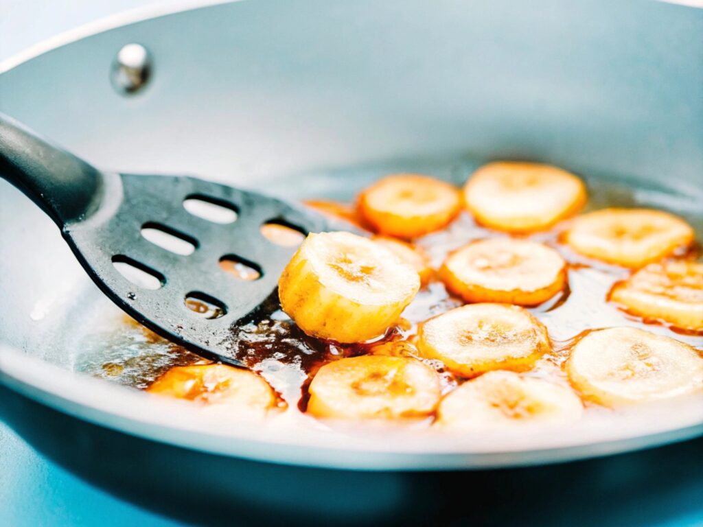 Banana slices frying in bubbling golden oil, with a slotted spoon lifting one slice out of the pan