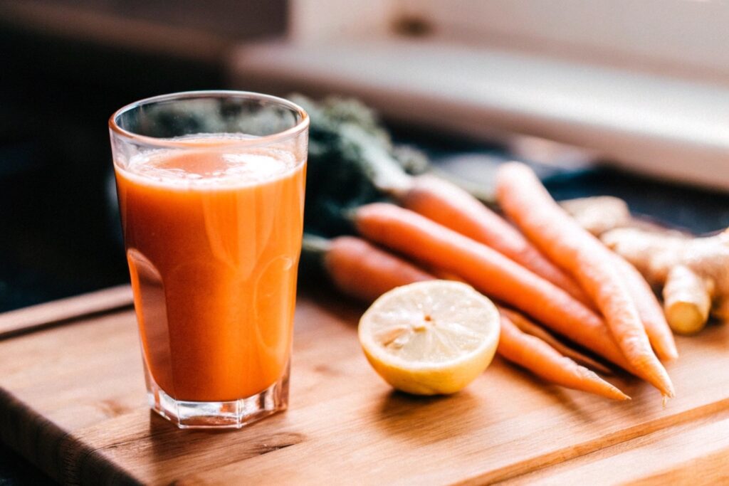 Close-up of a glass of carrot juice on a wooden counter, surrounded by fresh carrots, a lemon slice, and a small bowl of ginger slices, with warm natural lighting enhancing the scene