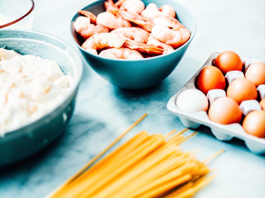 Ingredients for chicken and shrimp carbonara laid out on a marble kitchen counter, including Parmesan cheese, eggs, bacon, shrimp, and spaghetti