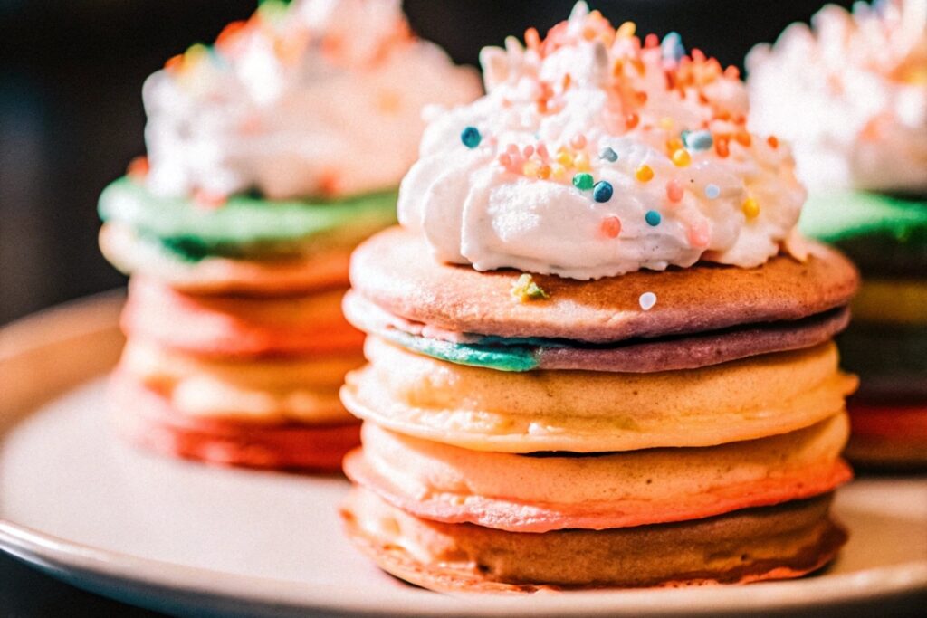 A colorful stack of rainbow hotcakes topped with whipped cream and sprinkles on a wooden table