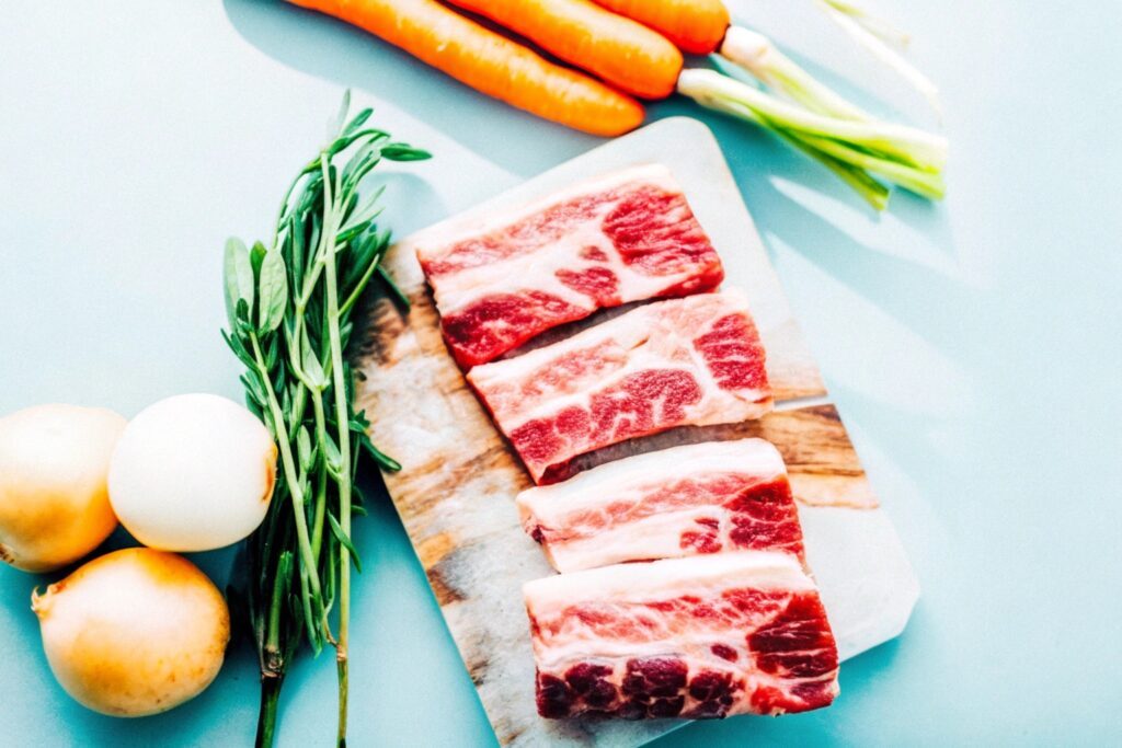 A flat-lay view of fresh ingredients, including boneless beef short ribs, vegetables, garlic, and herbs, arranged on a cutting board