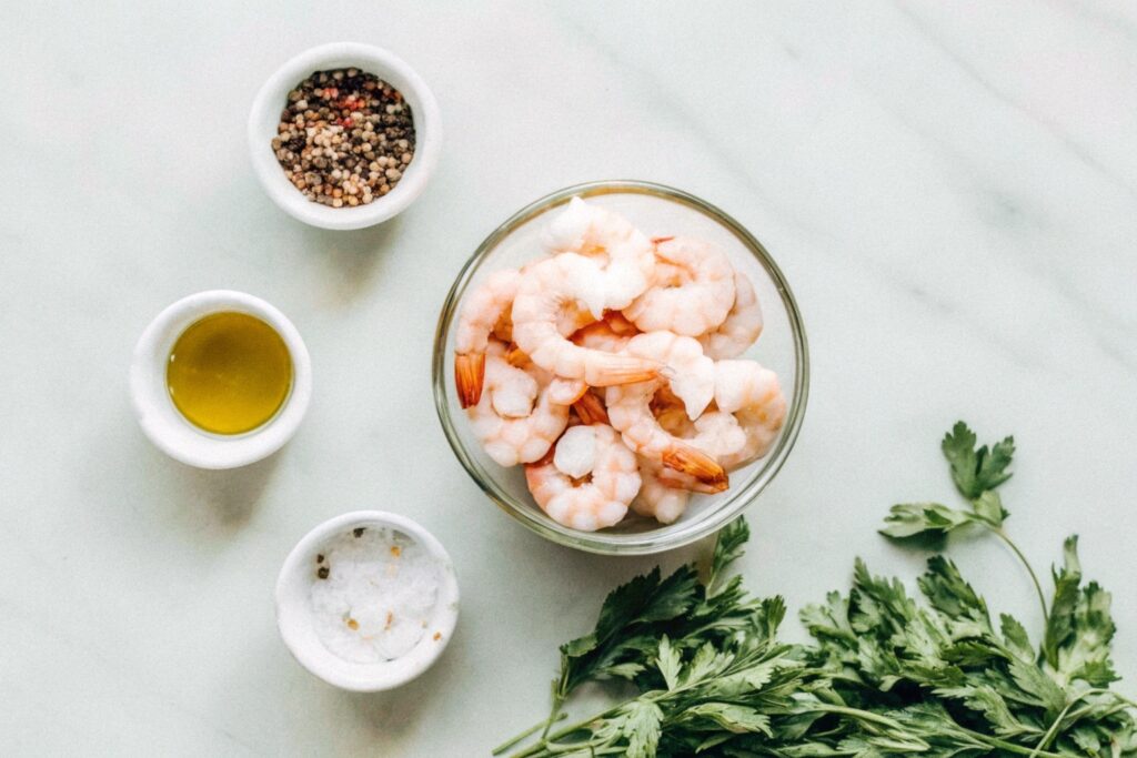 Raw shrimp in a bowl, surrounded by small dishes of spices, olive oil, and fresh parsley on a wooden countertop