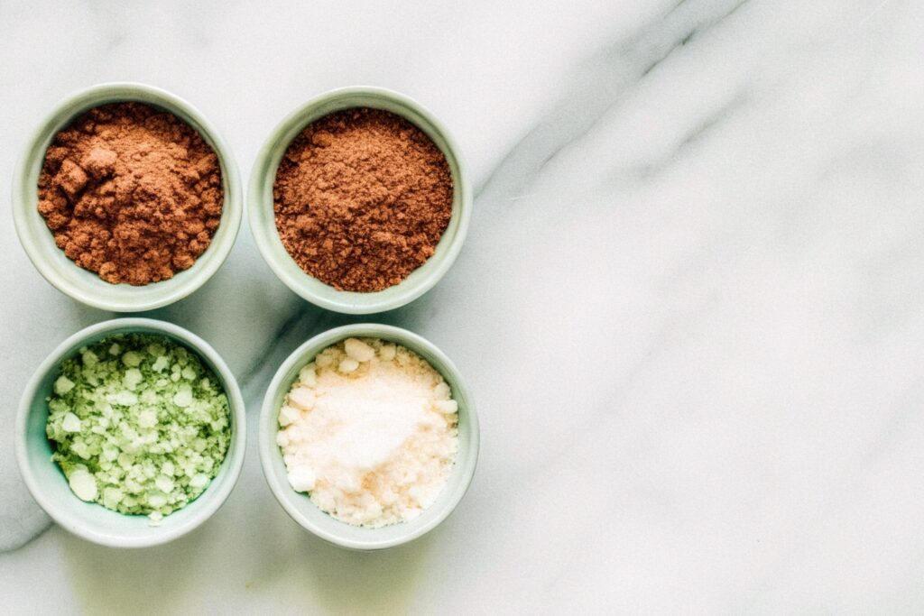 A flat lay of neatly measured ingredients in small bowls on a marble countertop, including red food coloring, cocoa powder, and butter