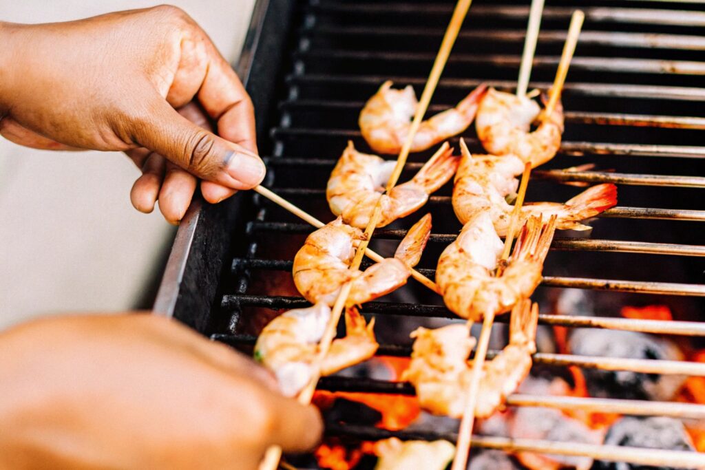 Hands flipping shrimp skewers on a grill with golden shrimp cooking over open flames