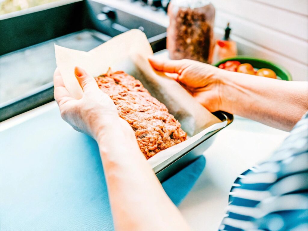 A person shaping the meat mixture into a loaf on a parchment-lined countertop, with spices and a smoker in the background