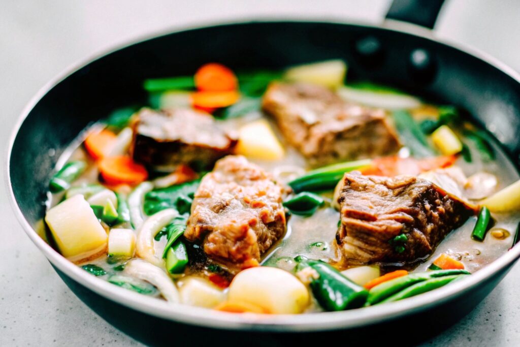 A skillet of boneless beef short ribs simmering in a thick, aromatic sauce with sliced carrots and celery visible