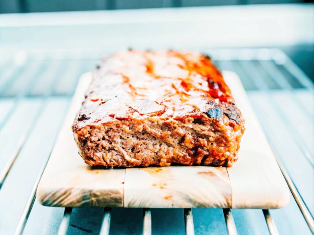 a bacon-wrapped smoked meatloaf on a wooden board, sliced to reveal a cheesy filling, surrounded by fresh herbs and sides

