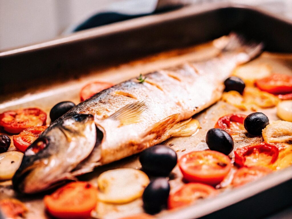 A whole branzino just out of the oven on a baking sheet, garnished with roasted tomatoes and olives