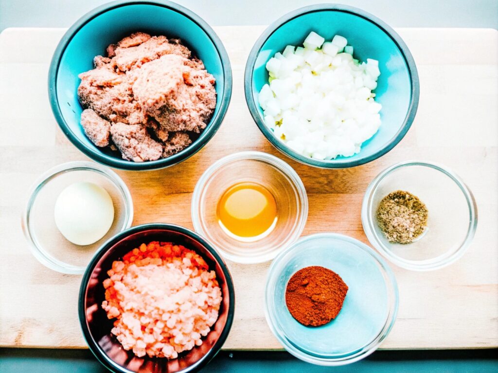 A wooden countertop displaying all the ingredients for smoked meatloaf, neatly arranged in bowls and measuring cups.