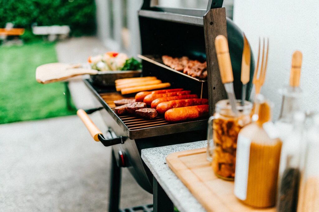 A backyard BBQ setup with a smoker releasing smoke, various meats on a grill nearby, and utensils and spices arranged on a table