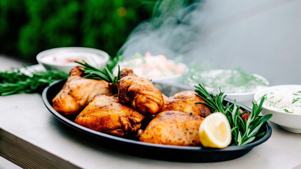 A platter of smoked chicken thighs with crispy golden-brown skin, garnished with fresh rosemary and lemon slices, surrounded by dipping sauces and coleslaw on a rustic wooden table