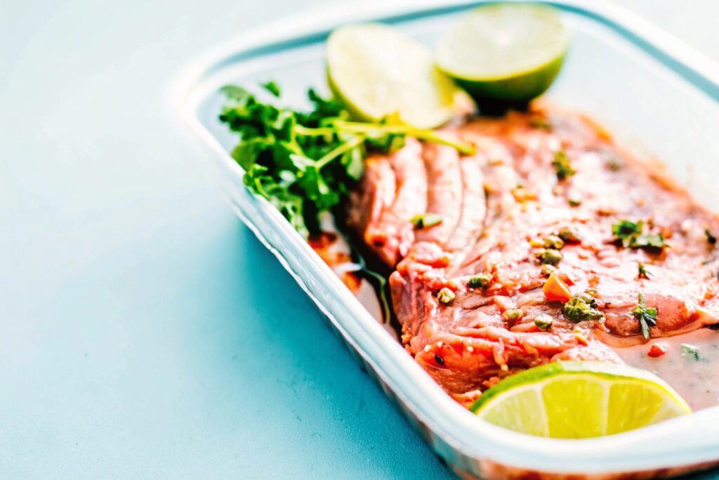 Marinated steak in a shallow dish, surrounded by lime wedges, garlic cloves, and fresh herbs, ready for grilling or searing