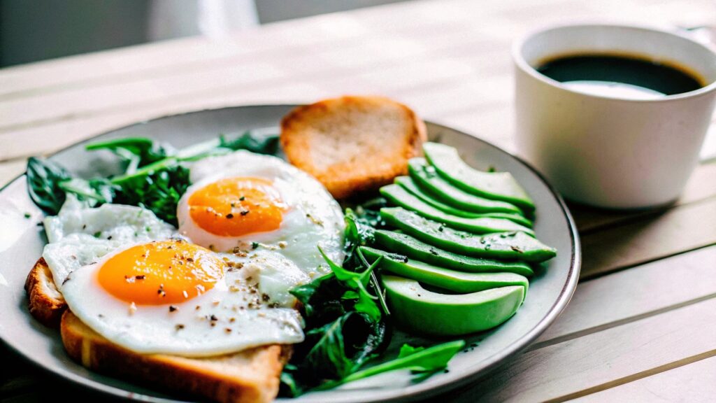 A beautifully plated breakfast featuring sunny-side-up eggs, avocado slices, and whole-grain toast, served on a wooden table with a cup of coffee nearby.