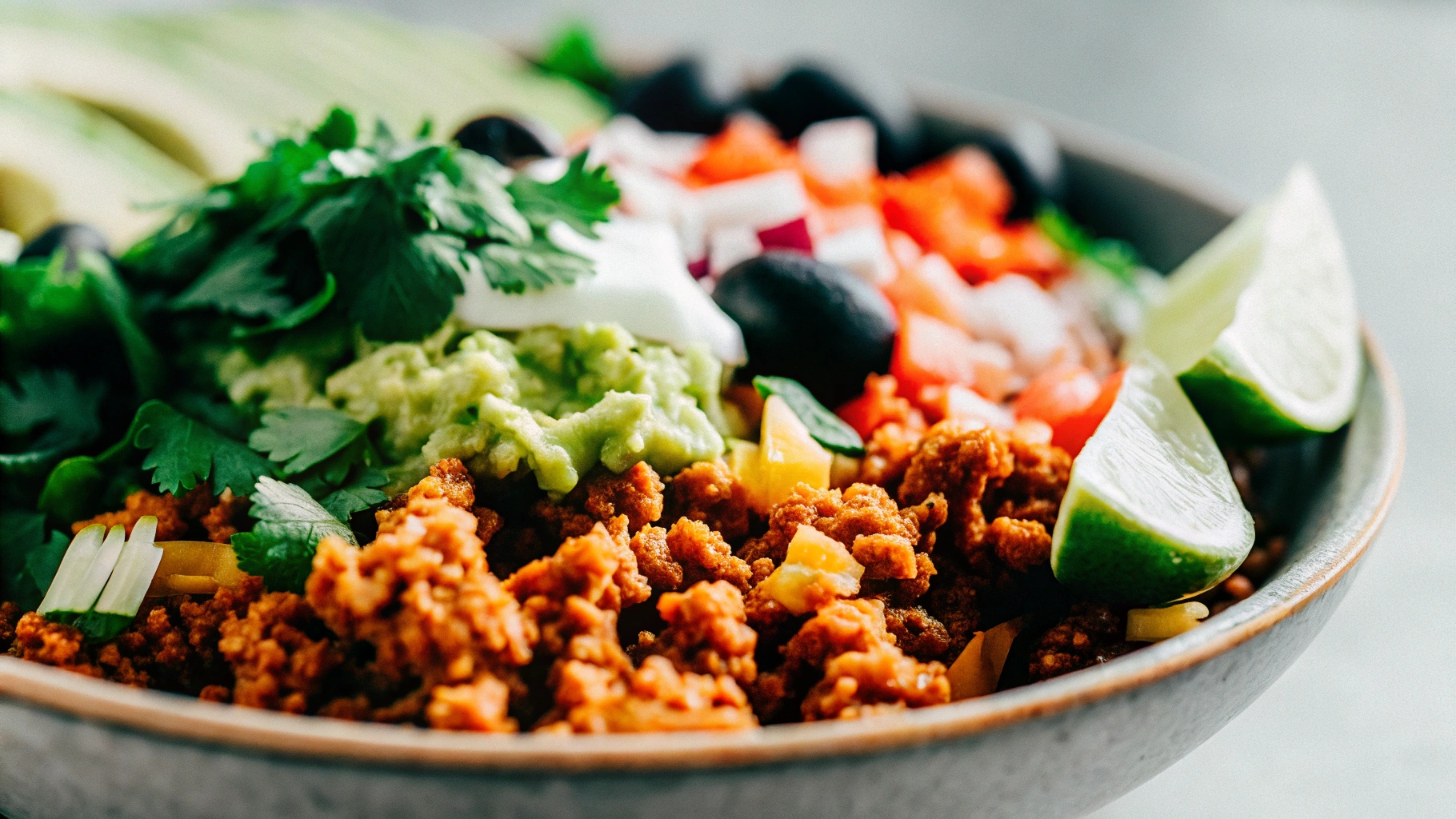 A close-up of a burrito bowl with Chipotle-style ground beef, topped with guacamole, salsa, and lime wedges, presented on a clean, modern surface