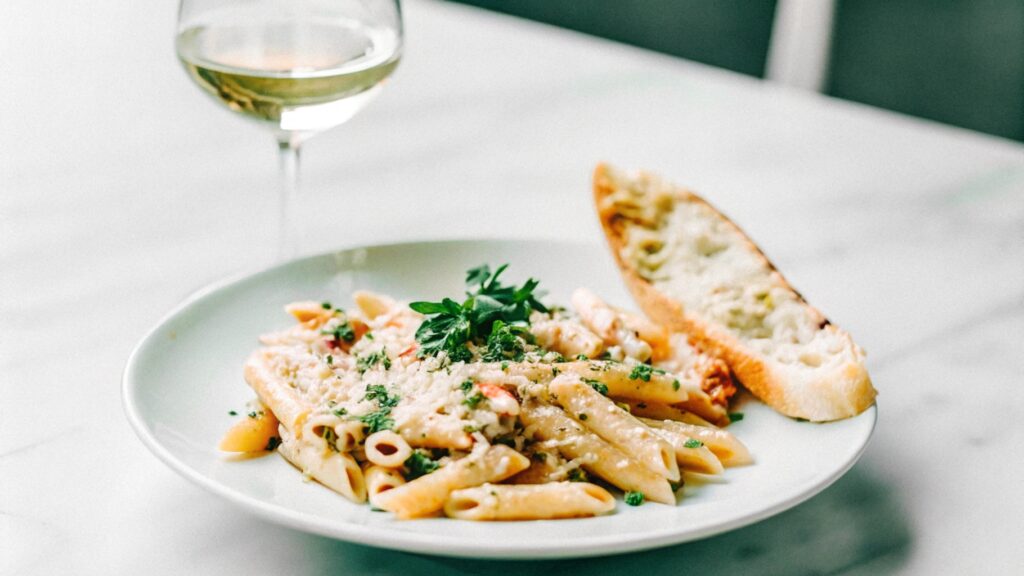 A beautifully plated serving of penne Alfredo, garnished with parsley, accompanied by crispy garlic bread and a glass of white wine on a neatly set dining table