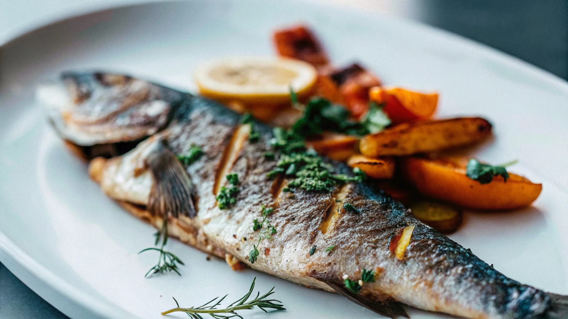 A whole roasted branzino on a white plate, garnished with fresh herbs, lemon slices, and a side of roasted vegetables