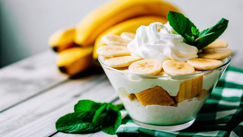 Classic banana pudding served in a clear bowl, featuring layers of creamy pudding, fresh banana slices, and golden wafers, garnished with whipped cream and a mint sprig.