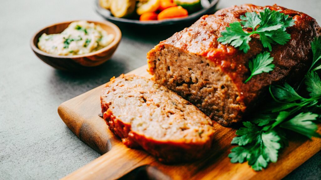 A beautifully sliced meatloaf on a rustic wooden cutting board, garnished with parsley, surrounded by side dishes like mashed potatoes and roasted veggies