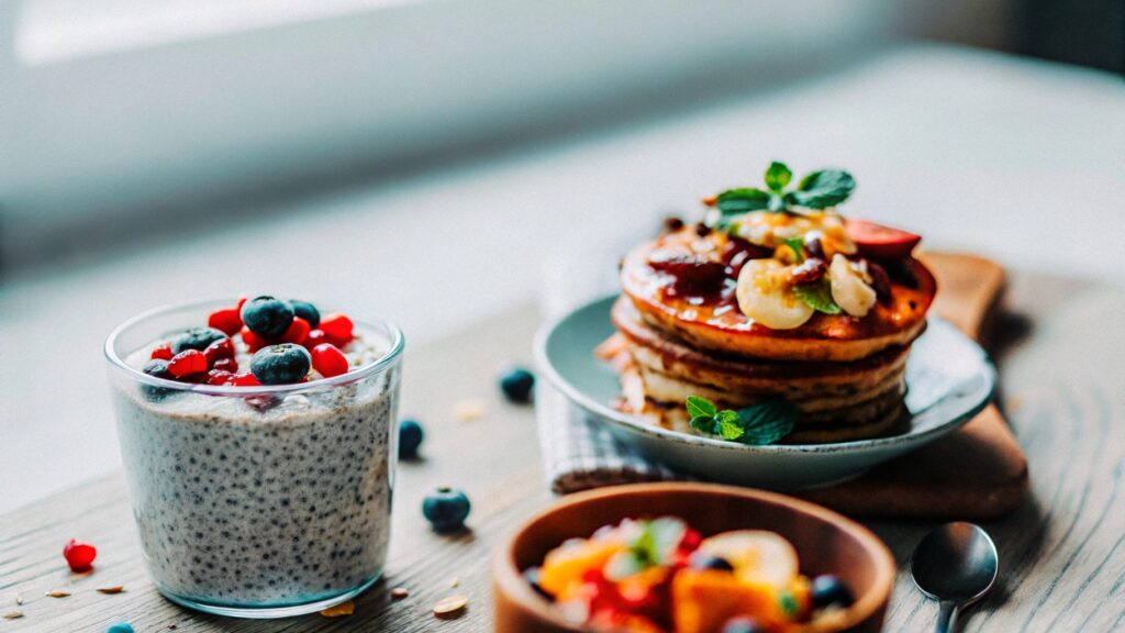 A rustic breakfast table displaying healthy sweet breakfast dishes: chia pudding with berries, banana oat pancakes with fresh fruit, and a tropical smoothie bowl, all arranged with natural light and garnished with fresh mint.