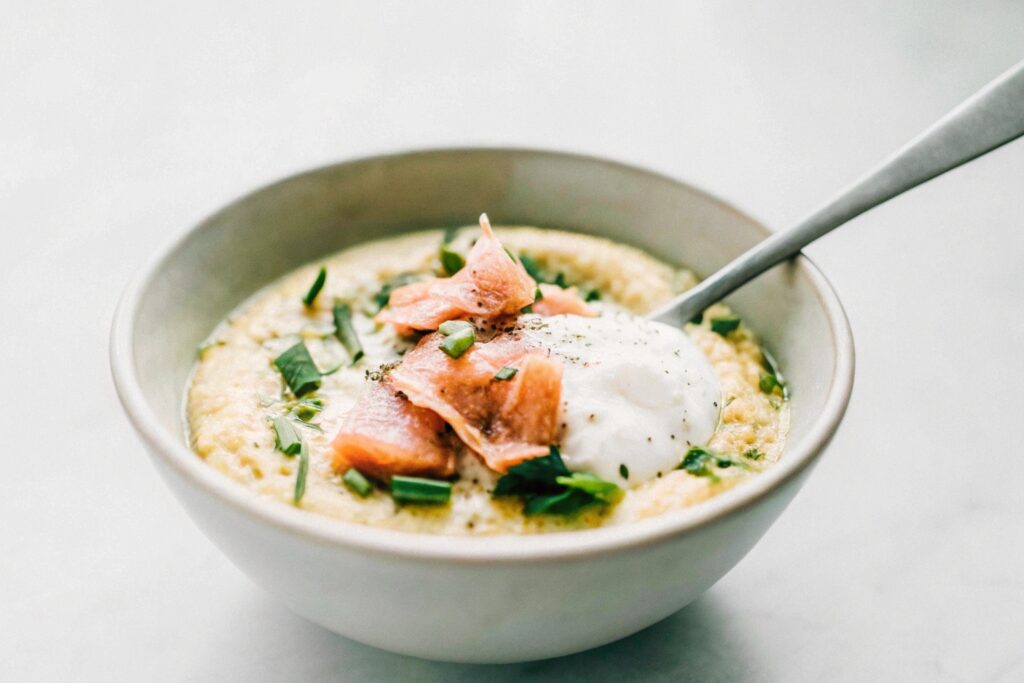 A bowl of creamy polenta topped with smoked salmon, crème fraîche, and fresh chives, served with a spoon