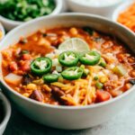 A bowl of taco soup surrounded by small bowls of toppings, including shredded cheese, jalapeños, lime wedges, and tortilla chips, on a wooden table