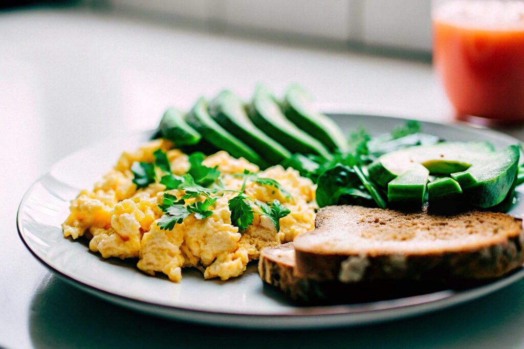 A breakfast plate featuring scrambled eggs, avocado slices, and whole-grain toast garnished with fresh herbs on a bright kitchen table