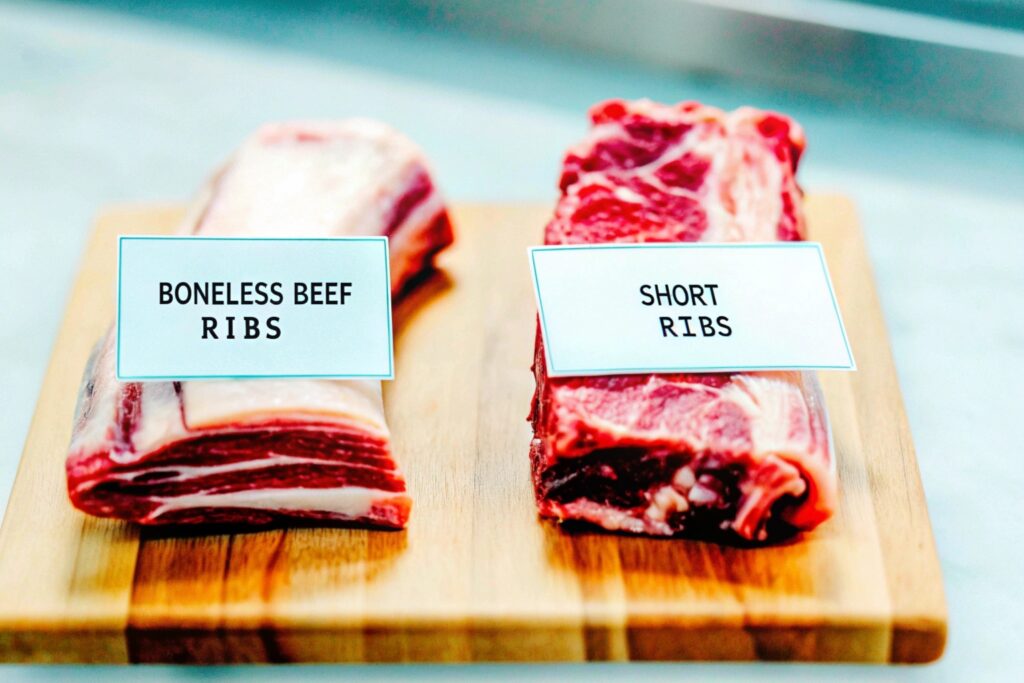 Boneless beef ribs and short ribs displayed side by side on a wooden cutting board, labeled to highlight their differences in cut and appearance