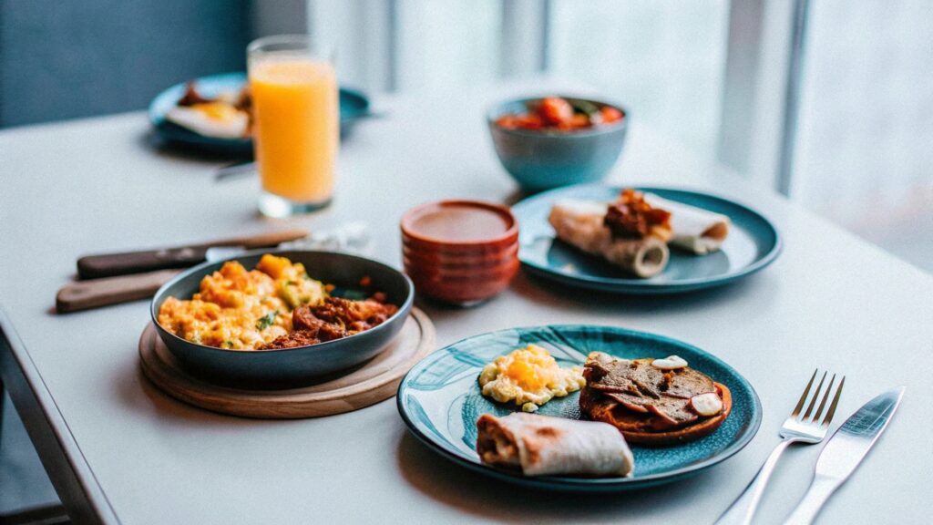 A cheerful breakfast table with several chicken dishes—scramble, wraps, and a sweet potato bowl—served alongside fresh orange juice and coffee