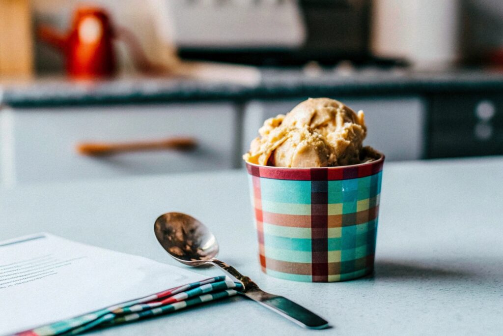 Cheerful kitchen scene with a tub of banana pudding ice cream, a serving spoon, and a handwritten note card with tips and FAQs next to the dessert.