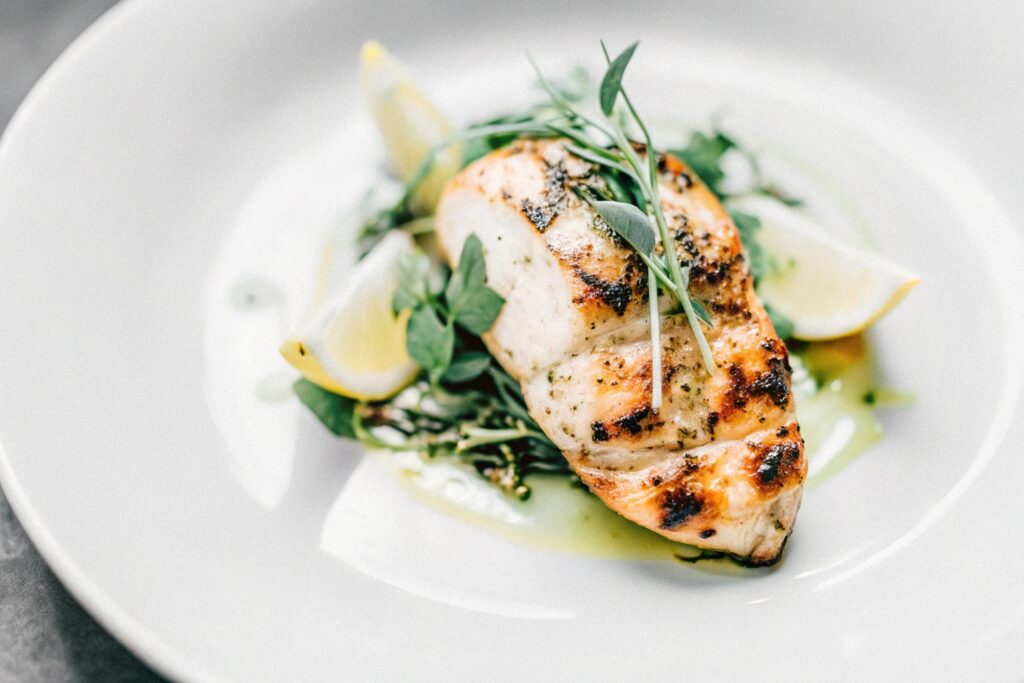 A close-up of a grilled chicken breast served with fresh herbs and lemon slices on a white plate.