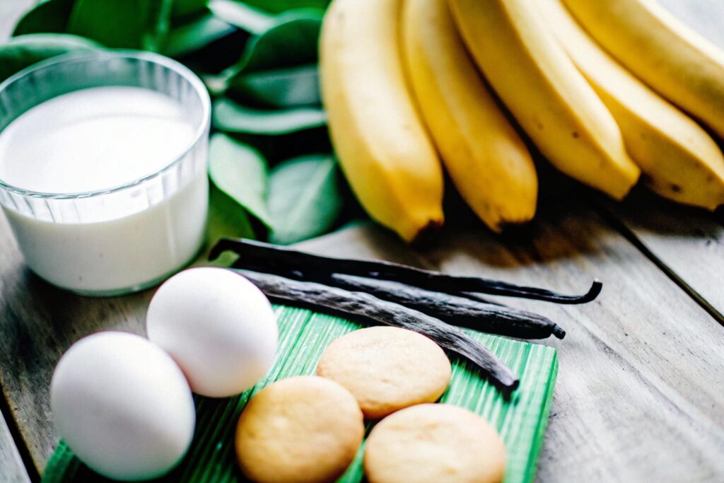 A close-up view of bananas, eggs, milk, and vanilla wafers arranged on a rustic wooden table, ready for making banana pudding