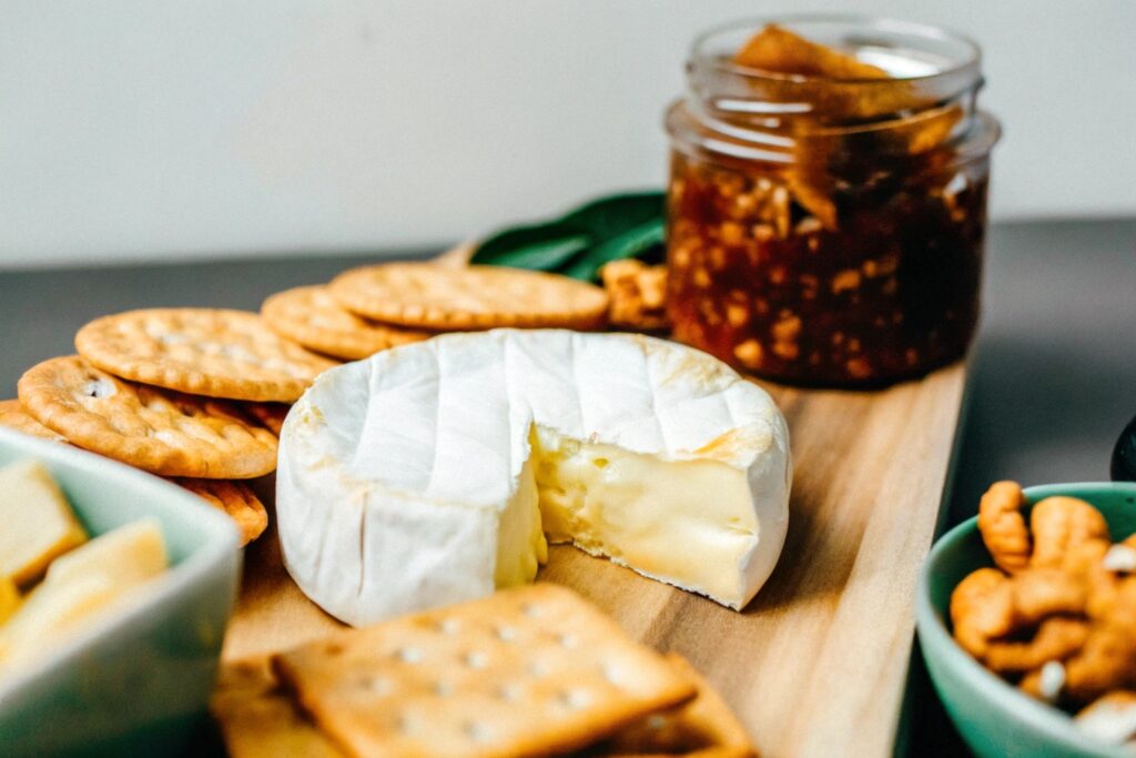 A close-up of Brie cheese on a wooden board surrounded by crackers, nuts, and a jar of fig jam