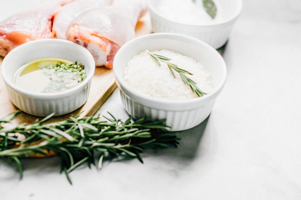 Close-up of chicken casserole ingredients: raw chicken, uncooked rice, fresh herbs, and a bowl of broth on a wooden board