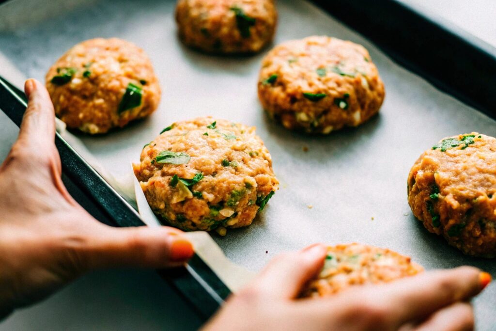 Hands shaping a salmon patty from the mixture, with several formed patties resting on a parchment-lined tray, ready for frying.