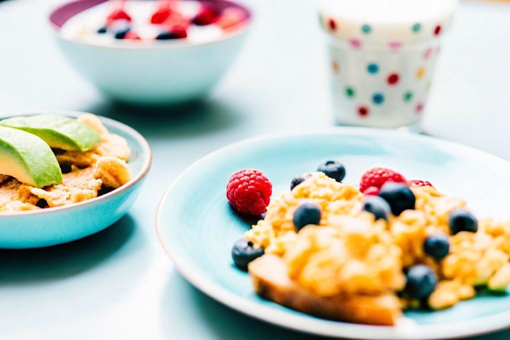 A breakfast table with scrambled eggs, avocado toast, and a bowl of whole-grain cereal topped with fresh berries and milk.