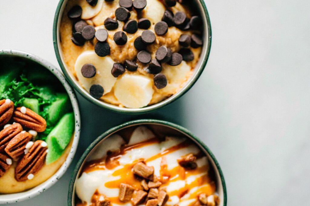 Three bowls of banana pudding ice cream with different toppings: chocolate chips, tropical garnishes, and pecans with caramel drizzle.