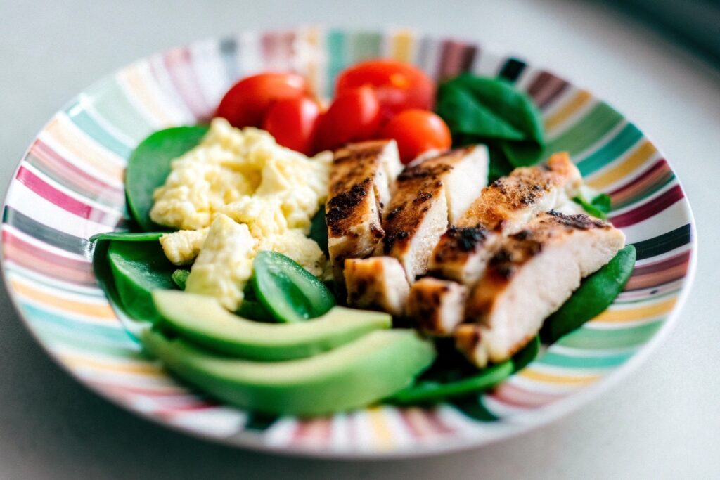 A colorful plate featuring grilled chicken breast slices served with scrambled eggs, avocado slices, and cherry tomatoes on a white plate