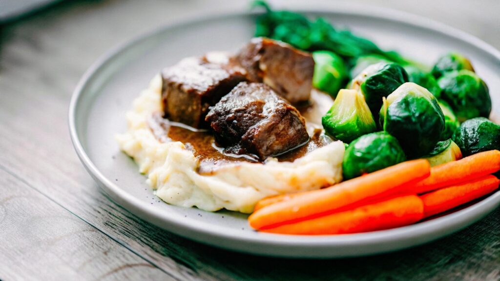 Boneless short ribs plated with creamy mashed potatoes, roasted carrots, and a drizzle of gravy, garnished with fresh herbs on a wooden table