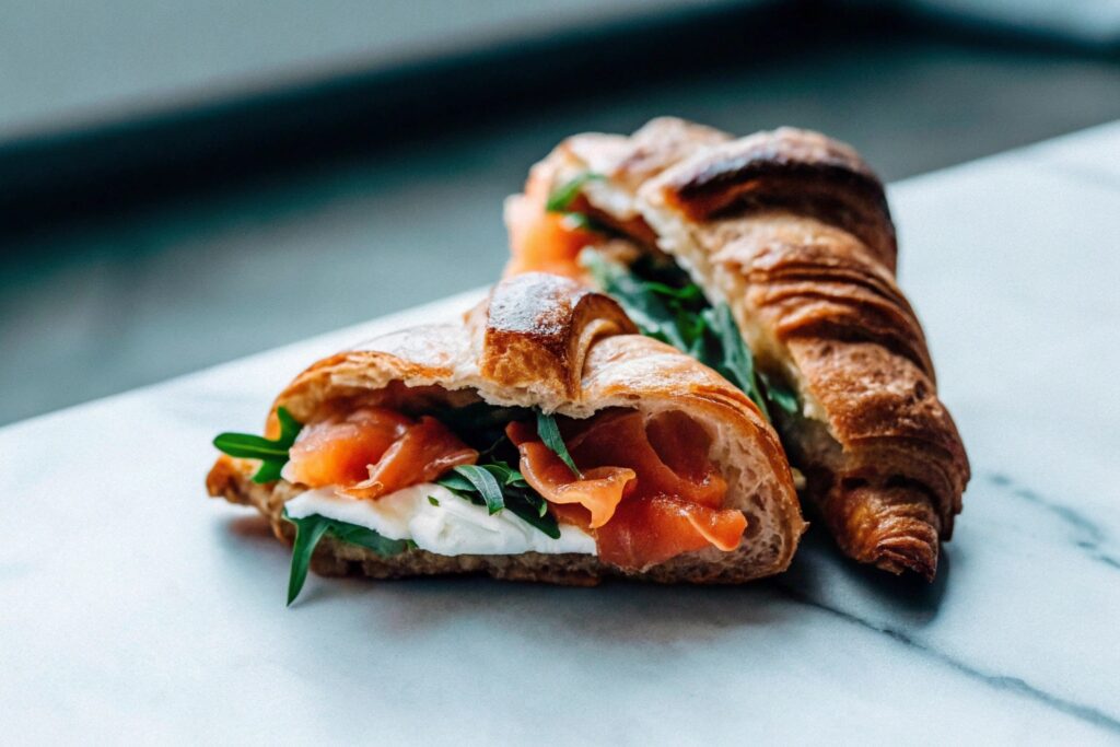 A golden croissant sliced open, filled with smoked salmon, cream cheese, and fresh arugula on a marble countertop