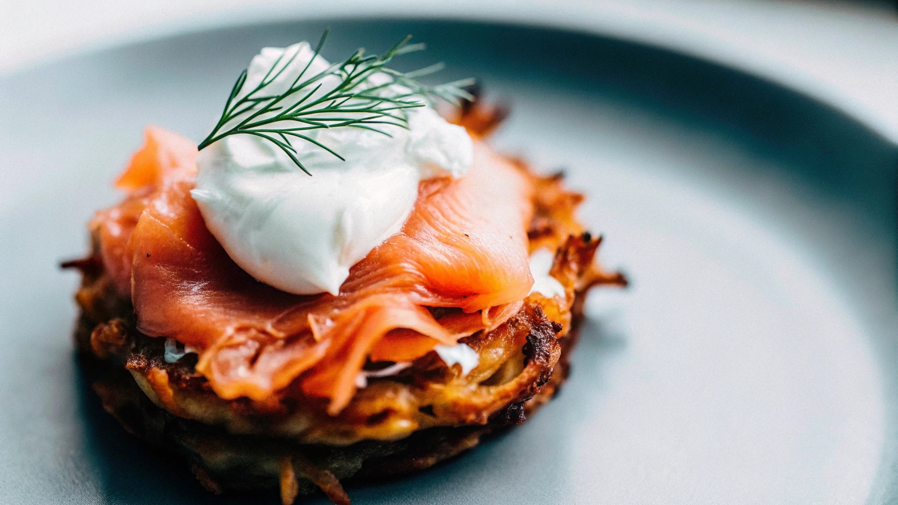 A golden potato rösti topped with smoked salmon and a dollop of sour cream, garnished with dill.